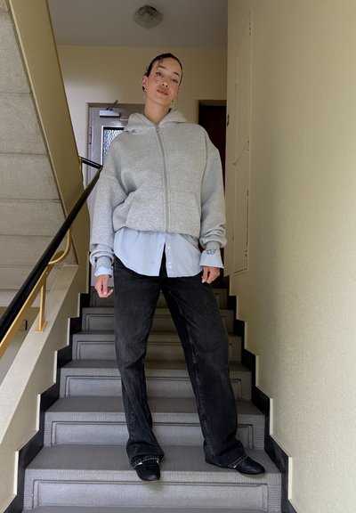 Sudadera gris con cremallera sobre una camisa de botones azul claro, combinada con pantalones vaqueros de pierna ancha oscuros y zapatos negros sin cordones, de pie en una escalera.