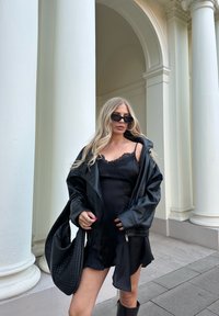 Czarna skórzana oversize'owa kurtka na bezrękawnikowej sukience z koronkowym wykończeniem, w połączeniu z czarnymi okularami przeciwsłonecznymi i torebką o fakturowanej powierzchni.