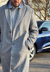 Homme portant un pardessus en chevrons gris et un pull se tient les mains dans les poches à côté d'une voiture bleue par une journée ensoleillée.