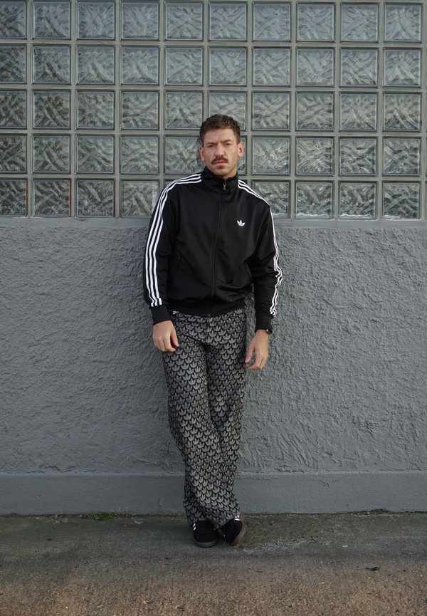 Άνδρας με μαύρο μπουφάν Adidas και παντελόνι με σχέδια, στηριζόμενος σε μια υφή γκρι τοίχου με γυάλινα μπλοκ από πάνω.