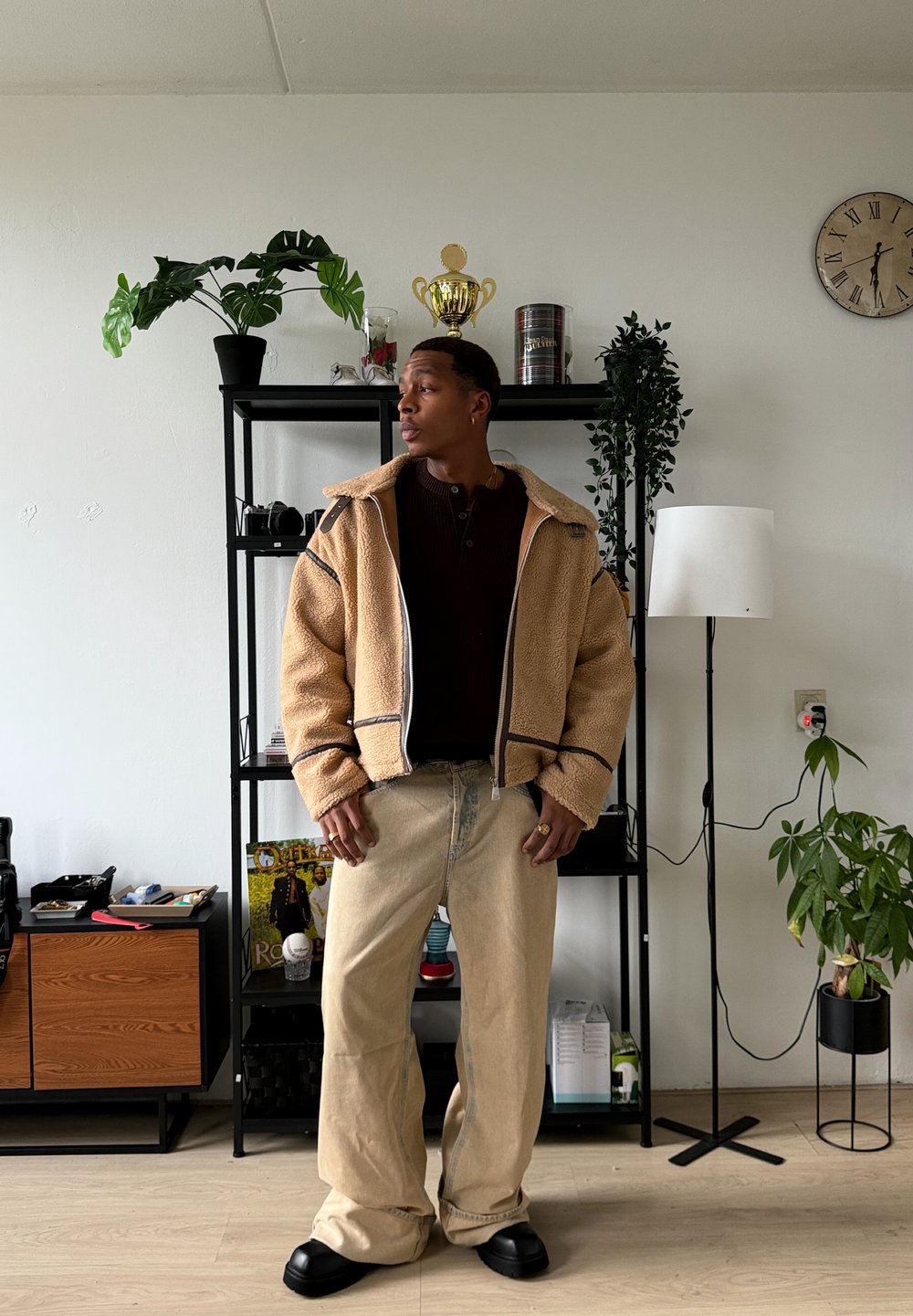 Veste en polaire beige avec une fermeture éclair, détails noirs, et un haut henley marron. Jeans larges de couleur claire et chaussures noires. Décor intérieur avec des plantes.