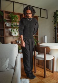 Suéter negro con pantalones acampanados y hombros redondeados, combinado con pantalones negros y zapatos negros, ambientado en un interior moderno lleno de plantas.