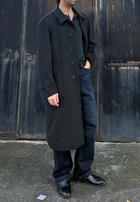 Άνδρας που στέκεται δίπλα σε φθαρμένη μεταλλική πόρτα, φοράει μακρύ σκούρο παλτό, μαύρο πουκάμισο, σκουρόχρωμα τζιν και μαύρα δερμάτινα παπούτσια, με το ένα χέρι στην τσέπη.