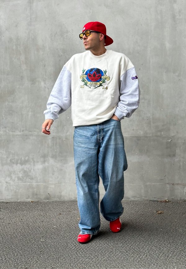 Sweatshirt oversized em branco e cinza com logótipo gráfico, emparelhado com jeans azuis folgados e sapatos vermelhos. Os acessórios incluem um boné vermelho e óculos de sol amarelos.