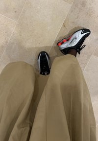 Osoba v béžových nohaviciach a čiernych, strieborných a červených Nike teniskách stojí na svetlej kamennej dlažbe.