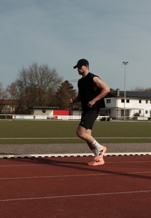 Un corredor con una camiseta sin mangas negra y pantalones cortos negros, con zapatillas de running en neón naranja, en una pista roja, frente a un fondo de campo verde.