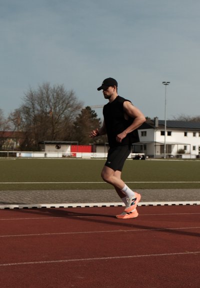 Un corredor con una camiseta sin mangas negra y pantalones cortos negros, con zapatillas de running en neón naranja, en una pista roja, frente a un fondo de campo verde.