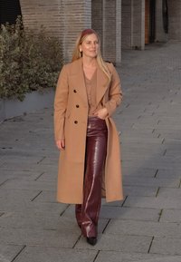 Dugi kamel kaput s crnim gumbima, složen preko bež majice. Uparen sa sjajnim burgundskim hlačama i špicastim crnim cipelama, na kamenitoj stazi.
