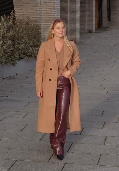 Abrigo largo de camel con botones negros, superpuesto a una blusa beige. Combinado con pantalones burdeos brillantes y zapatos negros de punta, sobre un camino de piedra.