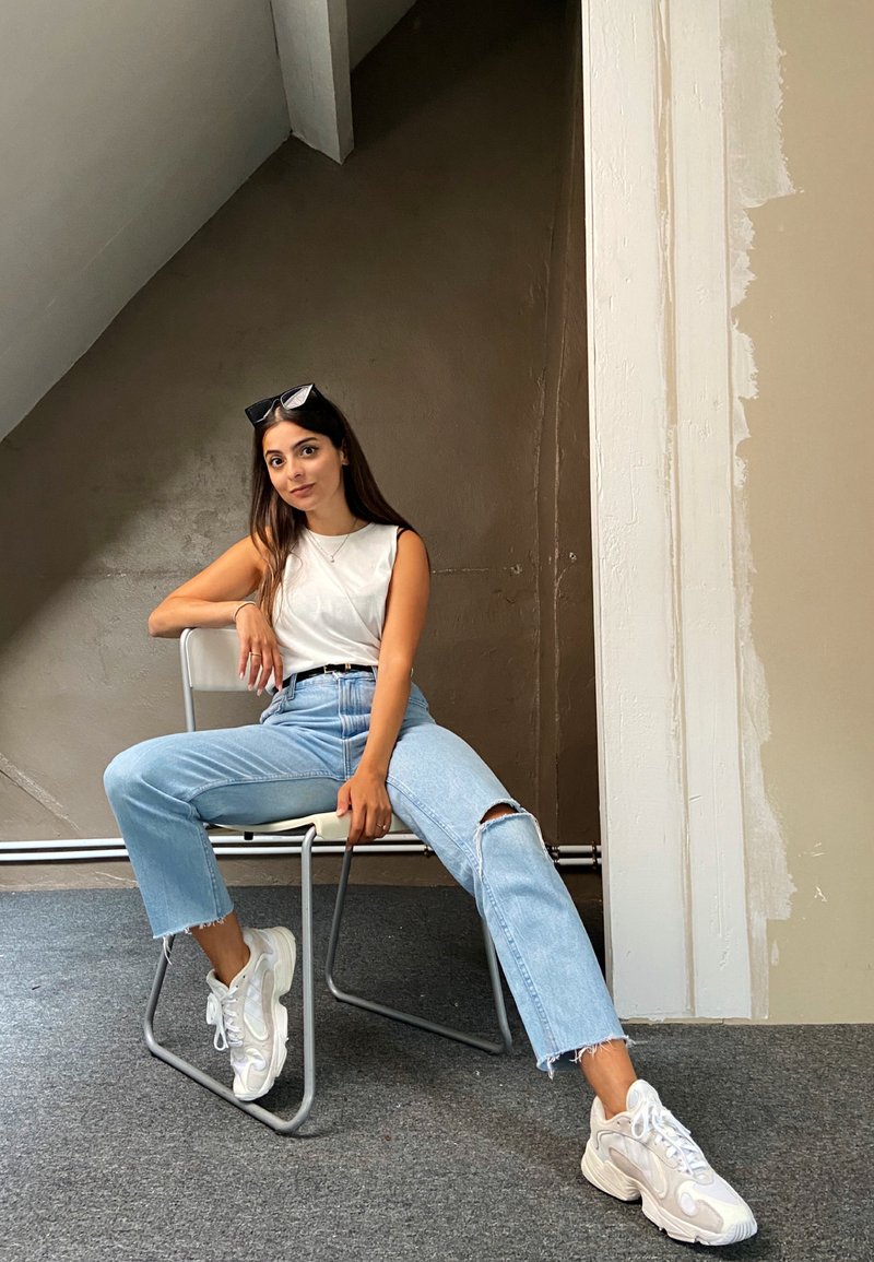 Világoskék, magas derekú farmernadrág, szakadozott szegéllyel és térdpírral, fehér ujjatlan felsővel és tömör fehér tornacipővel párosítva, fém széken ülve.