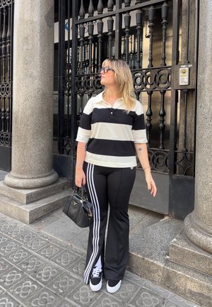 Mujer vestida con una camiseta de rayas blancas y negras y pantalones negros de Adidas con rayas blancas, sosteniendo un bolso negro junto a un pilar de piedra y una puerta de hierro forjado.