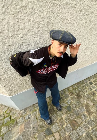 Jersey deportivo negro con acentos blancos, gorra de mezclilla azul y pantalones azules; accessorios con un collar de cuentas y zapatos negros sobre una superficie de adoquines.