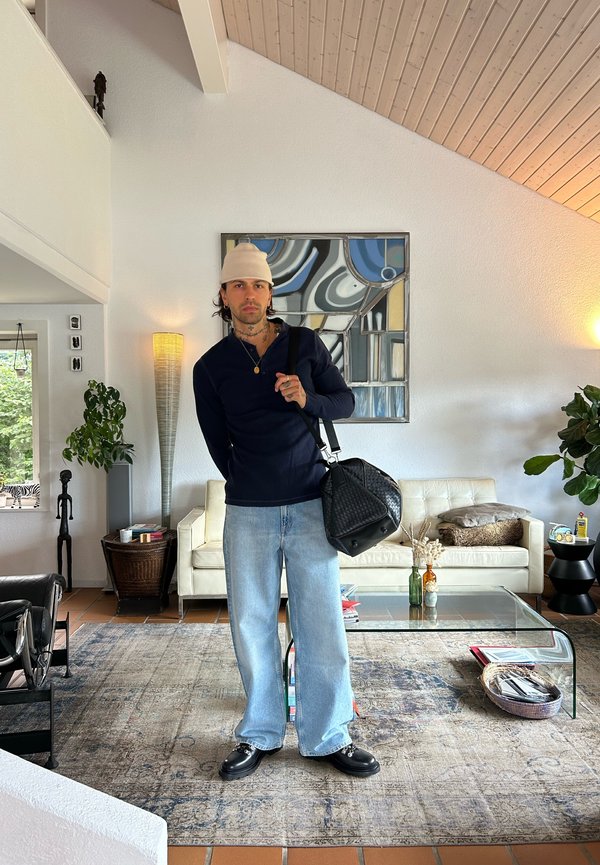 Man som bär en beige mössa, svart långärmad tröja, ljusblå jeans och svarta skor, står i ett vardagsrum och håller en svart väska.