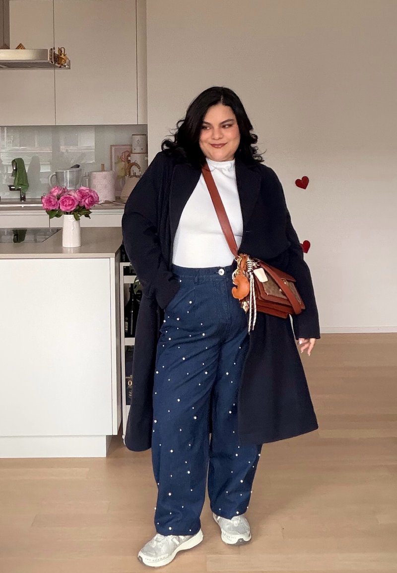 Femme portant un long manteau noir, un haut blanc, un pantalon bleu avec des détails en perles, des baskets blanches et un sac bandoulière marron dans une cuisine moderne.