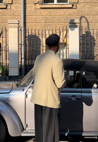Sivi vintage automobil je djelomično vidljiv, a pored njega stoji osoba u svijetloj prugastoj jakni i crnim hlačama, okrenuta prema van.