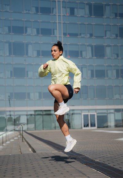 Chaqueta de pista ligera, amarilla neón, pantalones cortos negros y zapatillas de running blancas. Sujeto en medio de un salto en un entorno urbano con edificios de cristal.