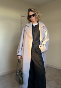 Femme portant un manteau gris clair surdimensionné, des lunettes de soleil noires, un pull sombre, un pantalon large noir, tenant un sac à main vert olive dans un cadre intérieur neutre.