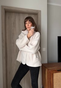 Νεαρή γυναίκα με λευκό φλις μπουφάν και μαύρο παντελόνι στέκεται μέσα σε εσωτερικό χώρο, αγγίζοντας το γιακά του μπουφάν της, με μια ξύλινη πόρτα και ντουλάπι στο φόντο.