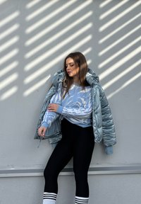 Light blue padded jacket with a shiny finish, layered over a blue and white patterned jumper, paired with black leggings and striped socks.