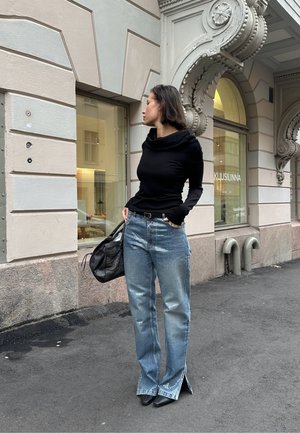 Schwarzes Langarmoberteil mit einem drapierten Ausschnitt, hellblaue gemusterte Jeans mit ausgestellt geschnittenen Säumen und eine schwarze Handtasche, kombiniert mit schwarzen Absätzen.