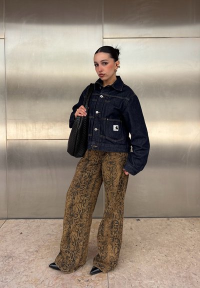 Joven con cabello oscuro recogido, que lleva una chaqueta vaquera negra, pantalones anchos marrones con estampado de serpiente, zapatos negros puntiagudos y sostiene un bolso negro.