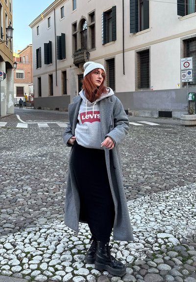 Abrigo largo gris sobre una sudadera gris de Levi's, combinado con una falda negra y botas de plataforma negras. El fondo presenta una calle de adoquines y edificios.