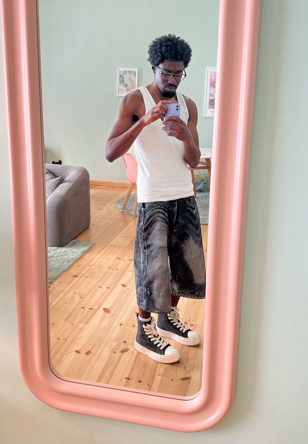 Schwarze und graue übergroße Jeansshorts, weißes Tanktop und schwarze High-Top-Sneaker mit weißen Sohlen, reflektiert in einem pinke gerahmten Spiegel.
