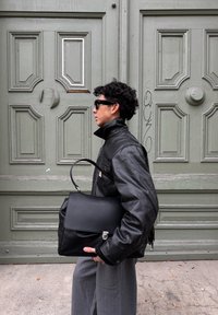 Melns ādas vēstneša soma ar pārsegu, sudraba detaļām un noņemamu plecu siksnu; persona melnā žaketē stāv pie zaļām durvīm.