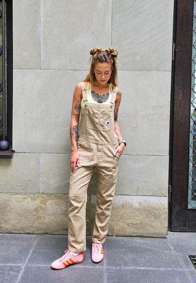 Pantalones de tirantes color beige con tirantes ajustables, bolsillo frontal y bolsillos laterales, combinados con zapatillas rosas con rayas naranjas. Textura de tela suave.