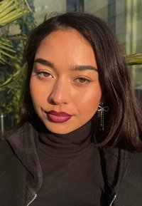 Porträt einer Person mit langen, dunklen Haaren, die einen schwarzen Rollkragenpullover trägt. Besonderheiten sind dunkellila Lippen und kreuzförmige Ohrringe.