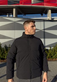 Μαύρο μπουφάν τύπου puffer με ψηλό γιακά, κλείσιμο με φερμουάρ και οριζόντια ραφή πάνελ. Φορεμένο πάνω από γκρίζα παντελόνια, μινιμαλιστικός σχεδιασμός, χωρίς ορατά λογότυπα.