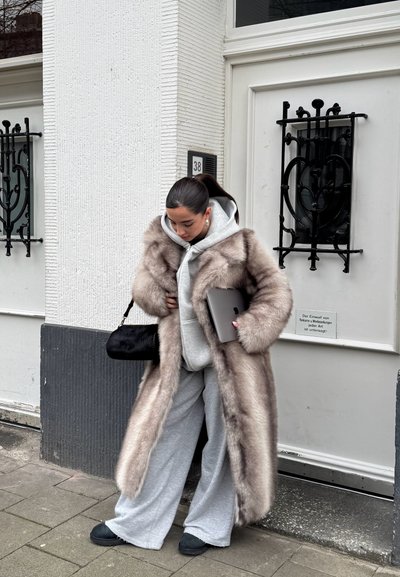 Abrigo largo de piel sintética en beige, sudadera gris debajo, pantalones de chándal grises, bolso negro al hombro y calzado negro, sosteniendo una tableta.