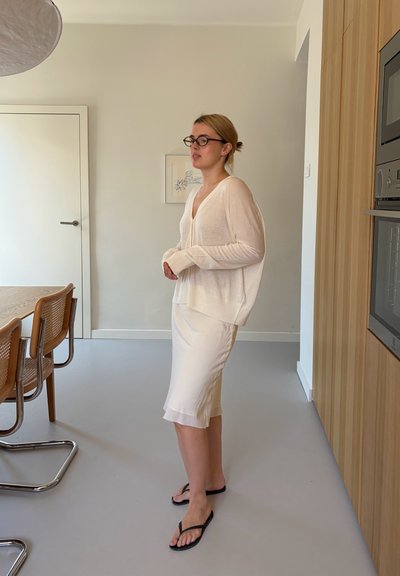 Mujer con gafas, blusa blanca y falda, de pie descalza sobre un suelo claro en una cocina y comedor modernos.