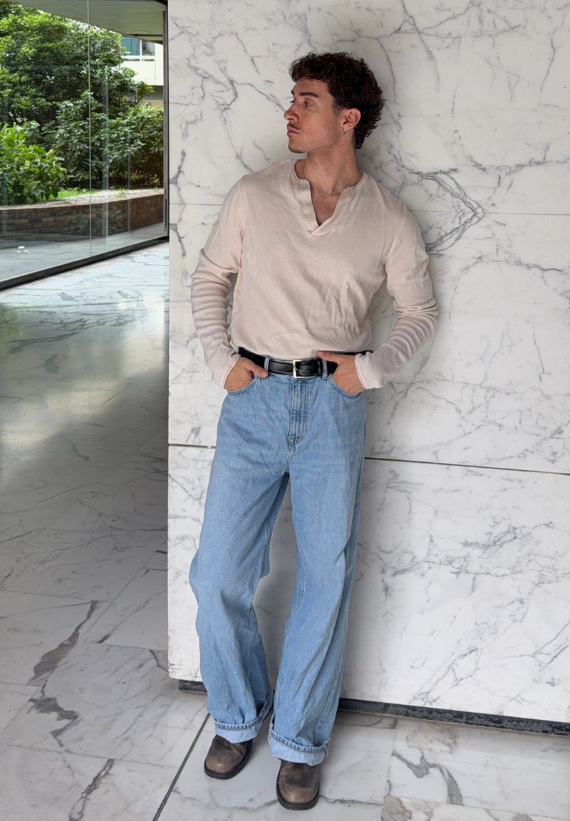 Man with curly hair stands against white marble wall wearing beige long sleeve shirt, light blue wide jeans, black belt, and brown shoes.