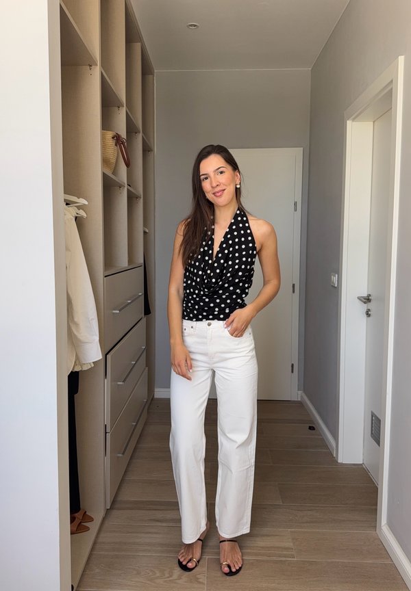 Frau steht in einem Flur und trägt ein schwarzes gepunktetes Neckholder-Oberteil, weiße Hose und schwarze Sandalen, lächelt in die Kamera.