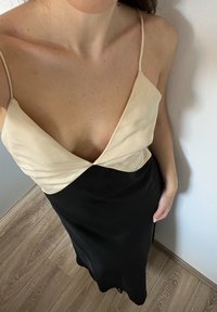Satin slipkjole med en cremefarvet top og sort nederdel; har en dyb V-udskæring og tynde, justerbare stropper. Glat tekstur og elegant design.