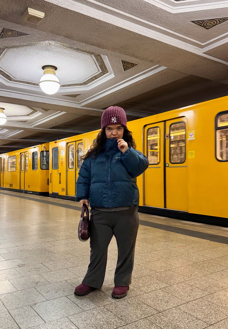 Tyrkysovo modrá zateplená bunda, sivá košeľa a dlhé kockované nohavice sú nosené s vínovou čiapkou a zodpovedajúcou obuvou, držiaci vínovú tašku. V pozadí je vidieť žltý vlak metra.