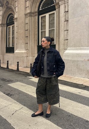 Mujer con cabello oscuro recogido en un moño, vestida con chaqueta de mezclilla, suéter gris, pantalones anchos de camuflaje y zapatos planos negros, de pie en un paso de peatones en una calle de la ciudad.