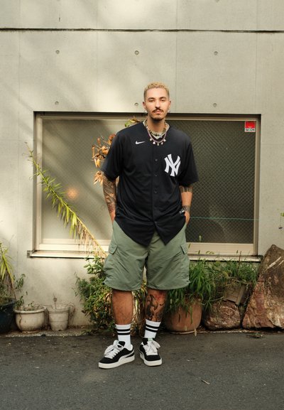 Jersey de béisbol negro de los NY Yankees, pantalones cortos de carga verdes, zapatillas blancas y negras, collares en capas, con macetas y una ventana de fondo.