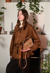 Brun, lager-på-lager-jacka med dragsko i midjan och tryckknappar; håller en brun handväska med grönt inslag; byxor med avslappnad passform.