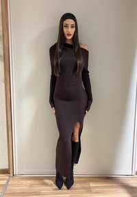 Une femme se tient contre un mur uni, portant une longue robe noire épaules dénudées avec une fente sur la cuisse, un serre-tête noir et des bottes noires pointues.