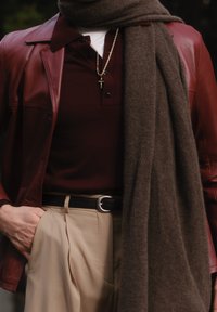 Bruine leren jas over een bordeauxrode polo, bruine wollen sjaal gedrapeerd, beige broek met een zwarte riem en een gouden ketting.
