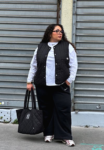 Chaleco acolchado negro sobre una camisa de manga larga blanca, combinado con pantalones de chándal negros. Sostiene un bolso de tote acolchado negro con un logo blanco.