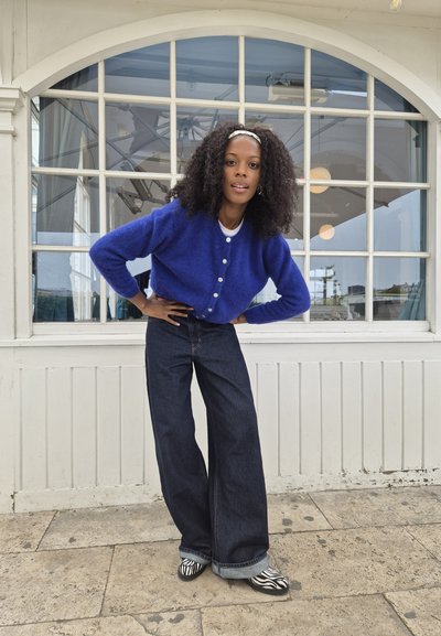 Cárdigan azul de felpa con cierre de botones, combinado con jeans de mezclilla oscuros de pierna ancha y zapatos de estampado de cebra. El fondo presenta grandes ventanales.