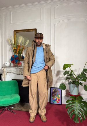 Hombre con chaqueta beige, suéter azul claro y gorra, de pie en una habitación con una silla verde, plantas decorativas, una obra de arte colorida enmarcada y un espejo ornamentado.