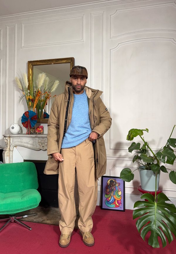 Homme en veste beige, pull bleu clair et casquette, debout dans une pièce avec une chaise verte, des plantes décoratives, une œuvre d'art colorée encadrée et un miroir orné.