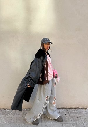 Persona joven con un abrigo largo negro sobre una camiseta rosa, pantalones deportivos grises rotos, gorra gris y zapatos gruesos grises, de pie contra una pared lisa.