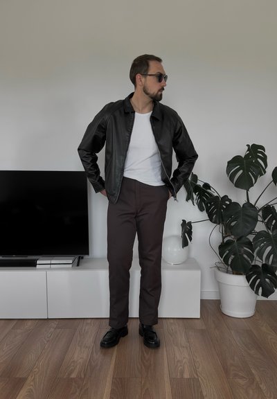 Chaqueta de cuero negra, camisa blanca ajustada, pantalones marrones, zapatos negros. Pose de pie, en una habitación con un televisor y una planta Monstera.