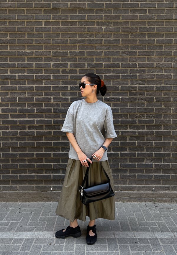 Frau mit Sonnenbrille, grauem Oversize-T-Shirt, olivgrüner weiter Hose, schwarzen Schuhen und einer schwarzen Handtasche, die vor einer dunklen Ziegelwand steht.
