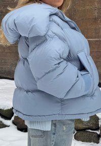 Femme portant une doudoune bleu clair les mains dans les poches, debout à l'extérieur sur un sol enneigé près d'un mur en pierre.